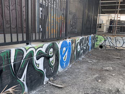 A concrete wall covered in colorful graffiti under a black metal fence, with dirt ground in the foreground and an industrial outdoor setting.