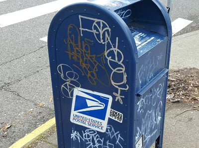A blue United States Postal Service mailbox on a street curb, covered with graffiti tags and stickers.